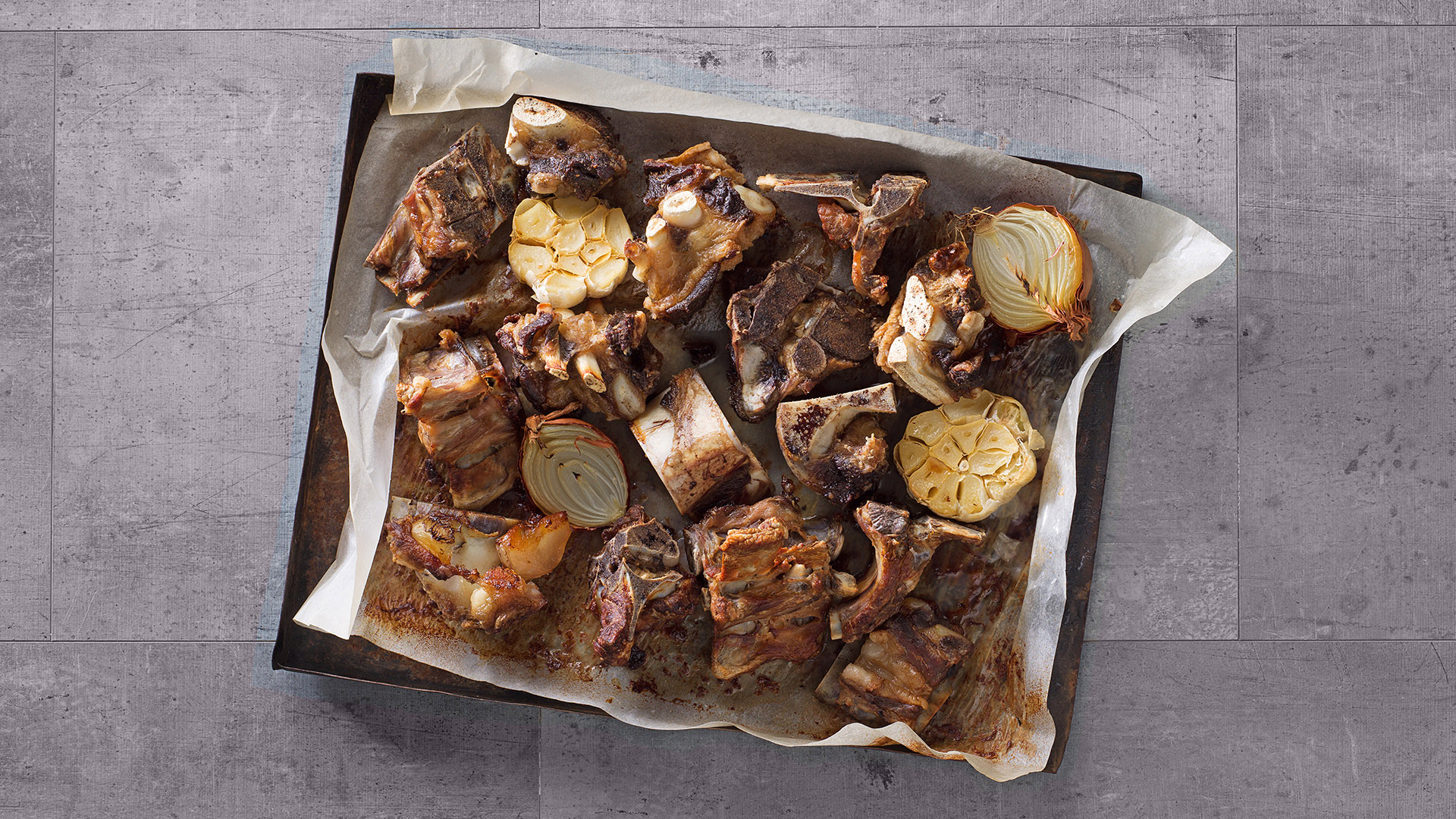 Diverse Knochen, Knoblauch und Zwiebel liegen braun gebacken in einem Backblech